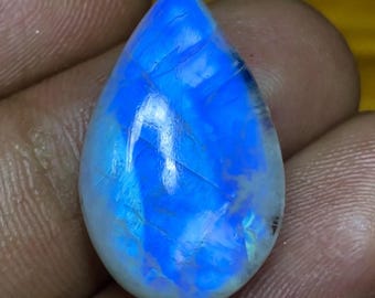 Könnte beinhalten: Ein polierter Mondstein in Tropfenform. Der Stein hat eine leuchtende, irisierende Qualität mit einer Mischung aus Blau- und Weißtönen. Der Edelstein wird in einer Hand gehalten und zeigt seine natürliche Schönheit und einzigartigen Farbvariationen.