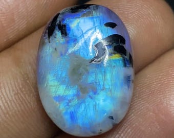 May include: Oval-shaped moonstone gemstone with a captivating display of blue iridescence, accented by black inclusions. The polished surface reflects light, creating a mesmerizing play of colors. A unique gemstone for jewelry.