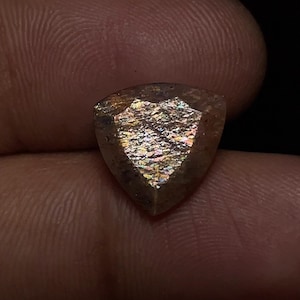 May include: A triangular-shaped gemstone with a shimmering, iridescent surface. The gem displays a range of colors, including gold, orange, and hints of green. The stone is held against a dark background, highlighting its unique facets and sparkle.