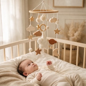 Può includere: Un mobile per bambini in legno con forme di stelle, nuvole e lune, insieme a elementi all'uncinetto, è appeso sopra un bambino in una culla. Il mobile presenta tonalità di legno naturale e accenti bianchi.