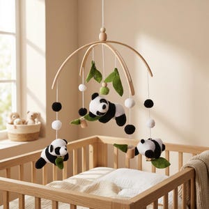 Op de afbeelding: Een hangende babymobiel met drie zwart-witte pluche pandaberen, opgehangen aan een gebogen licht houten frame. De mobiel bevat groene vilten bladeren, kleine houten kralen en afwisselende zwarte en witte pompons aan witte koordjes.