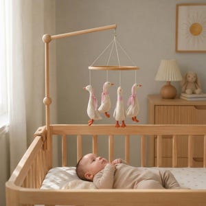 Pode incluir: Um berço de madeira com um móbile com quatro brinquedos de pelúcia de ganso branco com laços rosa. O móbile está pendurado sobre um bebê deitado no berço. O berço é feito de madeira clara.