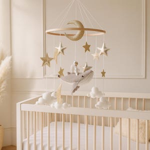May include: A baby mobile featuring a wooden ring with a gold glitter crescent moon and stars. Below, a gray whale carries an astronaut. White felt clouds and gold accents complete the celestial nursery decor.