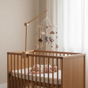 May include: A wooden cot with a hanging mobile featuring a koala, clouds, stars, and moons. The mobile is suspended above the cot, where a baby sleeps on a white mattress. The cot is made of light-coloured wood.