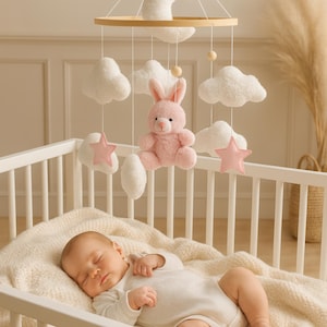 May include: A nursery mobile featuring a pink rabbit, white clouds, and pink stars suspended above a baby in a cot. The mobile hangs from a wooden ring. The baby, in a white onesie, sleeps on a cream blanket.