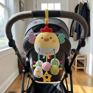 Pode incluir: Um brinquedo de pelúcia para carrinho de bebé em forma de galinha, com corpo amarelo, crista vermelha e asas estampadas. Pendura-se num clipe de plástico amarelo e inclui acessórios pendentes: um ovo branco com um pintainho e duas bolas texturizadas.