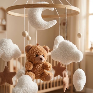 May include: A hanging mobile featuring a circular wooden frame with white plush clouds, a white plush crescent moon, brown felt stars, and a central brown plush teddy bear. The elements are suspended by white strings with small wooden beads.