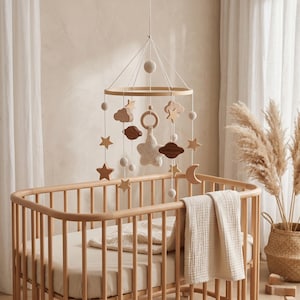 May include: A wooden baby mobile with star, cloud, and moon shapes hangs above a light-coloured cot. The mobile features a crochet star and wooden beads. A neutral-toned blanket rests inside the cot.