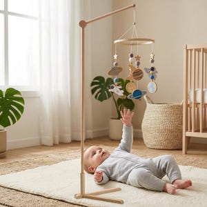 May include: A wooden baby mobile featuring space-themed elements including planets, stars, and astronauts suspended from a circular frame. The mobile is attached to a tall, light-colored wooden stand with a tripod base, designed for nursery use.