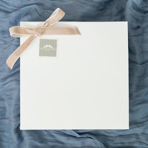 May include: A white square box with a beige velvet ribbon tied in a bow. The box has a small square label with text. The box rests on a blue fabric with a wrinkled texture.
