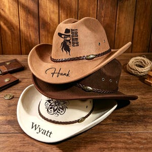 Può includere: Una pila di tre cappelli da cowboy in color cuoio, marrone scuro e crema. Il cappello color cuoio presenta una grafica con teschio nero con il testo COWBOY e il nome Hank. Il cappello crema mostra un disegno floreale e il nome Wyatt. Ogni cappello ha una fascia intrecciata.
