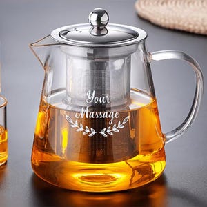 Pode incluir: Um bule de vidro transparente com tampa de aço inoxidável e um infusor de malha removível no interior. O corpo de vidro apresenta o texto impresso em branco Your Massage acima de um gráfico decorativo de folhas.