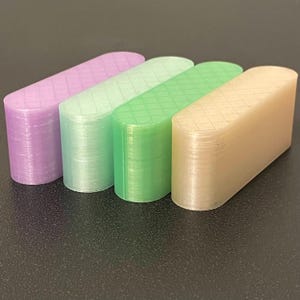 May include: Four elongated, pill-shaped objects with rounded ends and a flat top surface featuring a grid pattern. The objects are translucent and colored in shades of light purple, pale mint green, bright green, and a soft beige or light tan.