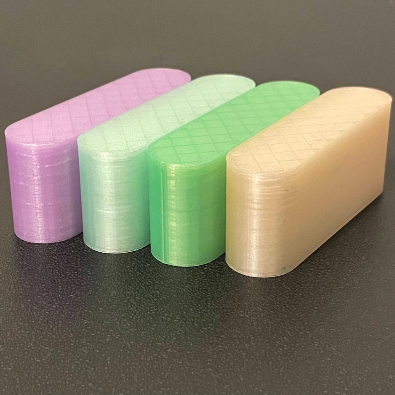May include: Four elongated, pill-shaped objects with rounded ends and a flat top surface featuring a grid pattern. The objects are translucent and colored in shades of light purple, pale mint green, bright green, and a soft beige or light tan.