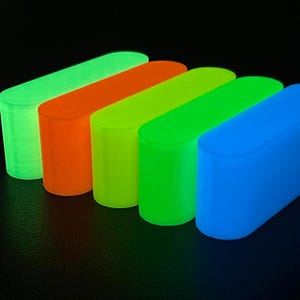 May include: Five rectangular blocks with rounded ends arranged in a row. The blocks are illuminated in vibrant, glowing colors including light green, orange, yellow-green, bright green, and blue, creating a luminous effect against a dark background.