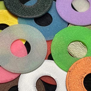May include: A collection of colorful, textured, circular objects with a hole in the center. The colors include pink, yellow, green, blue, purple, orange, white, and black. The objects appear to be made of a woven or fabric-like material.