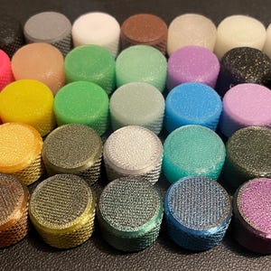 May include: A collection of small, cylindrical knobs arranged in a grid. Each knob features a textured, knurled side surface and a flat, circular top. The set includes a wide variety of solid, translucent, and glittery colors including red, orange, yellow, green, blue, purple, pink, black, white, gray, and metallic tones.