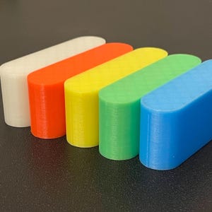 May include: Five elongated, pill-shaped blocks arranged in a row, featuring a smooth, matte finish. The colors from left to right are white, orange, yellow, green, and blue. Each block displays a subtle, diamond-patterned texture on the top surface.