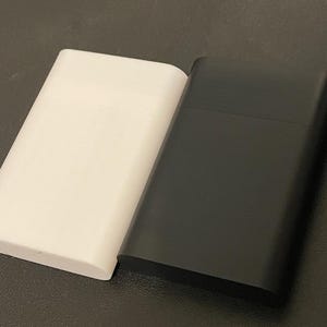 May include: Two rectangular cases, one in matte white and one in matte black, positioned side by side. Each case features a horizontal seam across the upper portion, indicating a lid or opening mechanism, with smooth, rounded edges and a flat surface.