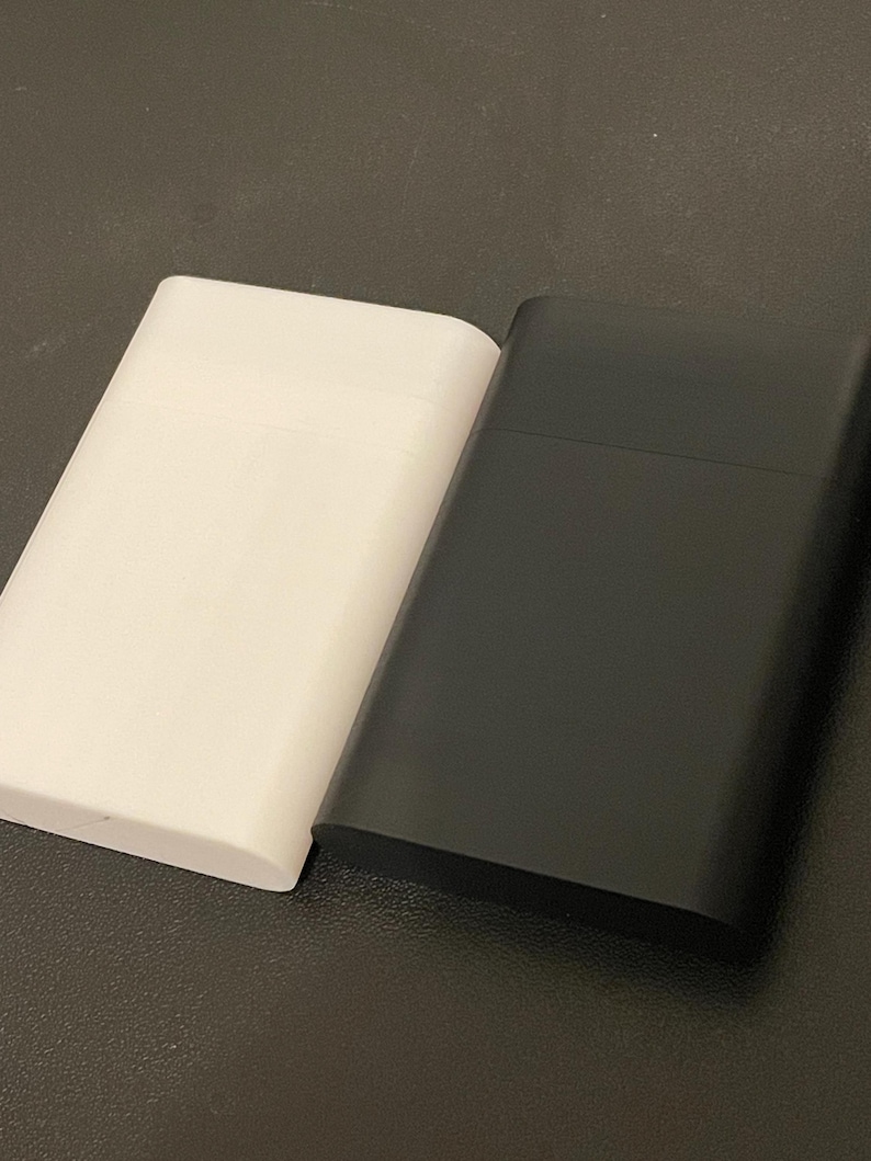 May include: Two rectangular cases, one in matte white and one in matte black, positioned side by side. Each case features a horizontal seam across the upper portion, indicating a lid or opening mechanism, with smooth, rounded edges and a flat surface.