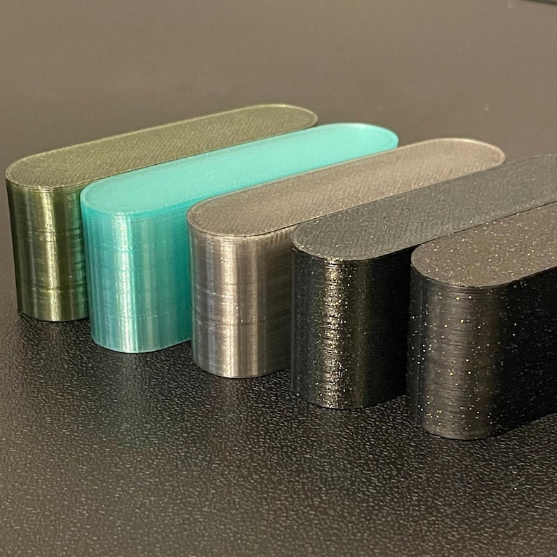 May include: Five elongated, pill-shaped objects arranged in a row. The colors include metallic olive green, translucent teal, metallic silver, matte black, and black with subtle silver glitter flecks. Each piece has a smooth, flat top surface.