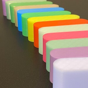 May include: A collection of elongated, oval-shaped blocks arranged in a diagonal row. The blocks feature a smooth, matte finish and come in a vibrant spectrum of colors including lavender, light green, beige, blue, lime, yellow, orange, cream, pink, and mint.