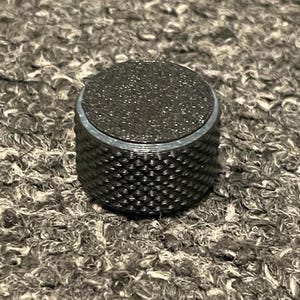 May include: A cylindrical metal control knob featuring a dark gray finish with a textured, diamond-patterned knurled side grip and a flat, matte black circular top surface. The component is designed for precise manual adjustment on electronic equipment.
