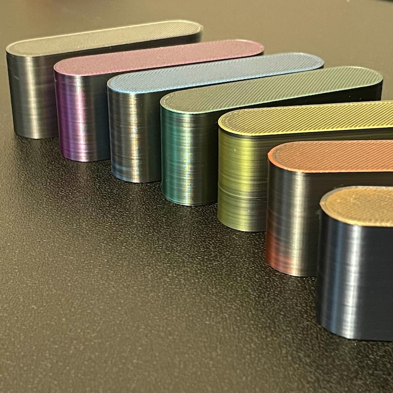 May include: A collection of seven elongated, oval-shaped blocks arranged in a diagonal row. Each block features a distinct metallic or iridescent finish, including shades of black, purple, blue, green, yellow, copper, and bronze with visible 3D print lines.