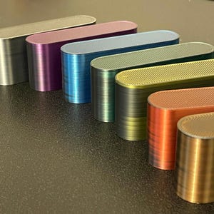 May include: A collection of seven elongated, oval-shaped blocks arranged in a diagonal row. Each block features a distinct metallic finish in silver, purple, blue, teal, green, orange, and gold, with a visible diagonal textured pattern on top.