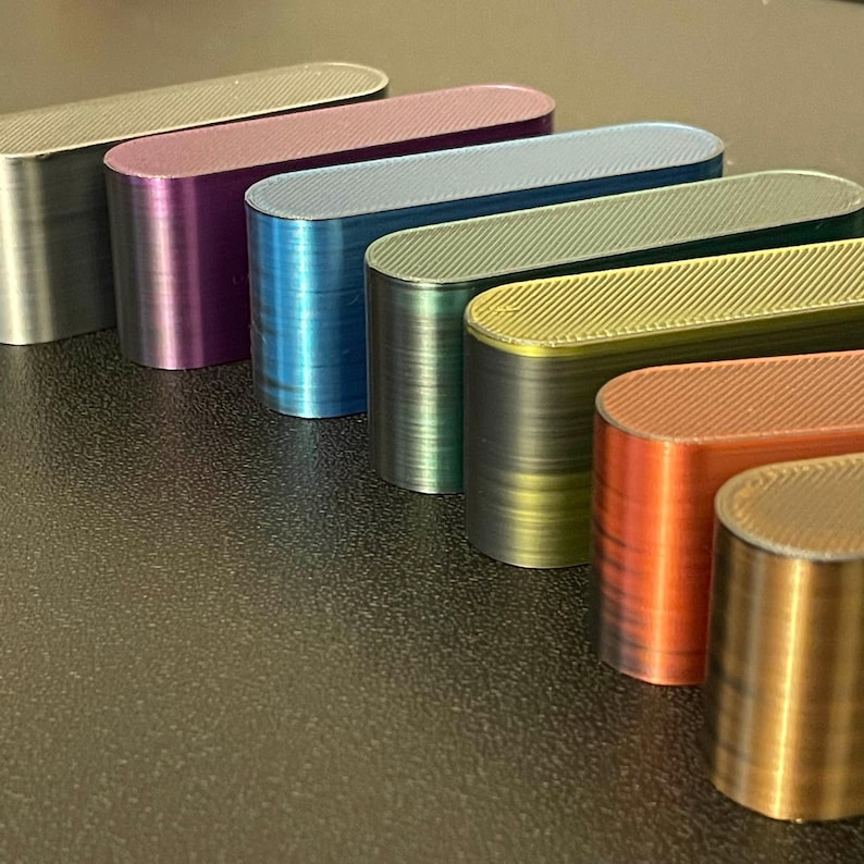 May include: A collection of seven elongated, oval-shaped blocks arranged in a diagonal row. Each block features a distinct metallic finish in silver, purple, blue, teal, green, orange, and gold, with a visible diagonal textured pattern on top.