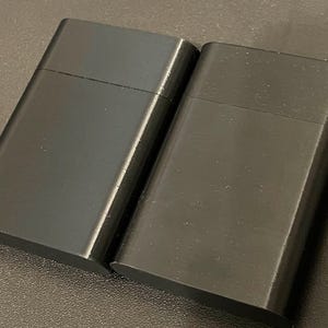 May include: Two rectangular black lighters with a smooth, matte finish. Each lighter features a horizontal seam across the upper portion, indicating a hinged lid design. The objects are positioned side-by-side, displaying a uniform dark color.