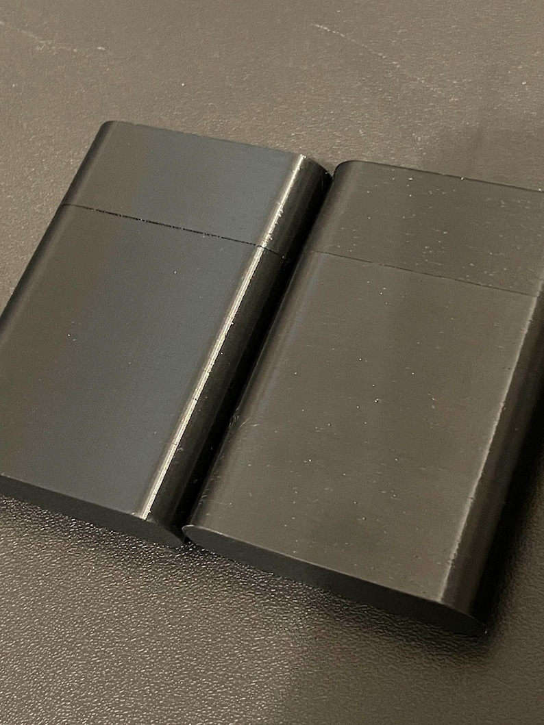 May include: Two rectangular black lighters with a smooth, matte finish. Each lighter features a horizontal seam across the upper portion, indicating a hinged lid design. The objects are positioned side-by-side, displaying a uniform dark color.