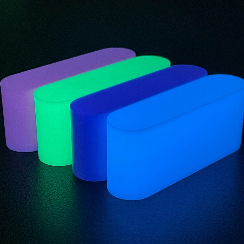 May include: Four rectangular blocks with rounded ends, each emitting a distinct glow. The colors from back to front are purple, bright green, dark blue, and light blue. These solid, smooth-surfaced objects are arranged in a parallel row.