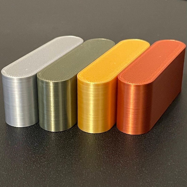 May include: Four elongated, pill-shaped blocks arranged in a row. The blocks are colored silver, olive green, gold, and copper. Each piece features a smooth, rounded top surface and vertical sides with a visible layered texture from 3D printing.