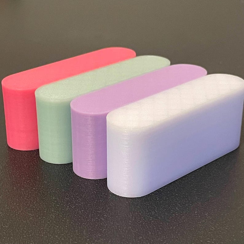 May include: Four elongated, pill-shaped blocks arranged in a row. The colors from back to front are vibrant pink, soft mint green, light lavender, and a pale, translucent white with a subtle diamond-patterned texture on the top surface.