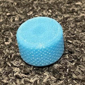 May include: A cylindrical knob in a bright light blue color. The top surface is smooth and flat, while the vertical side features a textured, diamond-patterned knurled grip. The object has a solid, uniform appearance with a matte finish.