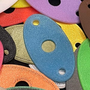 May include: A collection of colorful, oval-shaped 3D-printed objects with a hole in the center and a smaller hole at one end. The objects are in various colors, including blue, pink, orange, green, yellow, and purple. The objects have a textured surface.