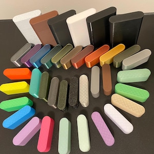 May include: A collection of rectangular, rounded-edge cases and smaller, elongated blocks in a wide variety of solid colors and textures. The items are arranged in a fan-like pattern, featuring matte, metallic, and textured finishes in shades of black, white, gray, brown, orange, yellow, green, blue, pink, and purple.