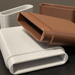 May include: A collection of three rectangular, hollow, open-ended sleeves with rounded corners. Two sleeves are light gray with a speckled texture, and one sleeve is solid brown. Each piece features a distinct horizontal ridge along the exterior.