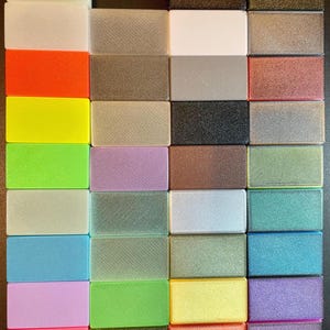 May include: A collection of thirty-two rectangular blocks arranged in a grid, featuring a diverse array of solid, metallic, and translucent finishes in colours including white, orange, yellow, green, blue, pink, black, silver, gold, and purple.