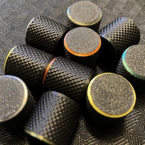 May include: A collection of black thimbles with textured surfaces and colorful accents. The thimbles have a diamond pattern and are accented with silver, yellow, orange, and green rims. The thimbles are scattered on a dark, patterned surface.