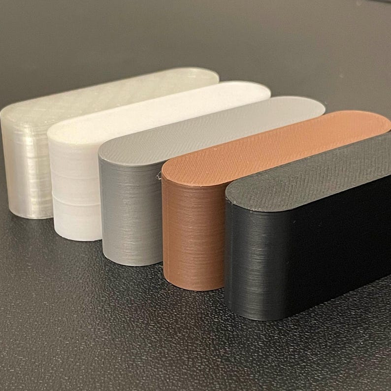 May include: Five elongated, oval-shaped blocks arranged in a row, featuring a smooth, layered texture. The colors include a translucent clear, solid white, light gray, warm brown, and solid black, all displaying a consistent 3D printed finish.