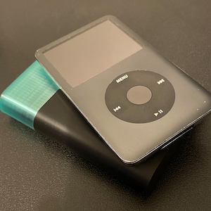 May include: A grey digital music player with a circular navigation wheel featuring MENU, play, pause, and track skip icons, resting on top of a black rectangular power bank that has a translucent teal-colored top section.