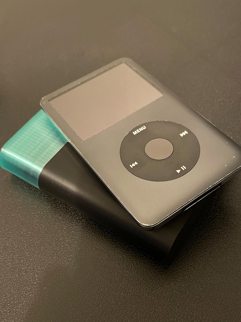 May include: A grey digital music player with a circular navigation wheel featuring MENU, play, pause, and track skip icons, resting on top of a black rectangular power bank that has a translucent teal-colored top section.