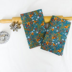 May include: Two teal tea towels with a floral pattern featuring orange, brown, and white leaves and flowers. The towels are folded and placed on a wooden rolling pin.