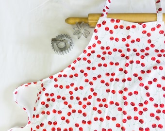 May include: White apron with a red cherry print. The apron has adjustable straps and a ruffled bottom.