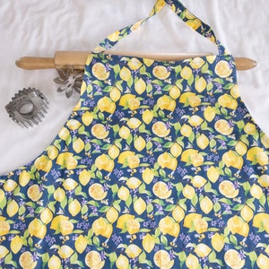 May include: A blue apron with a lemon print. The apron has yellow lemons, green leaves, and purple flowers on a dark blue background.