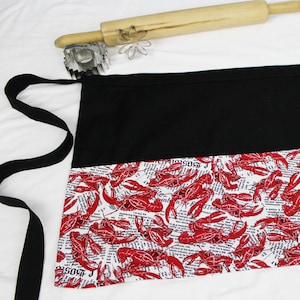 May include: Black and white apron with a red crawfish print. The apron has a black top section and a white bottom section with a red crawfish print. The apron has a black tie.