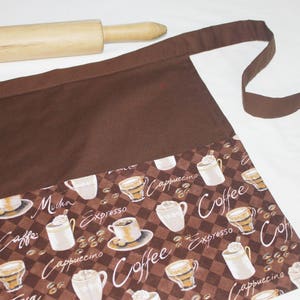 May include: Brown apron with a large pocket and adjustable straps. The pocket has a coffee themed print with images of coffee cups and the words "Coffee", "Cappuccino", "Espresso", and "Mocha".