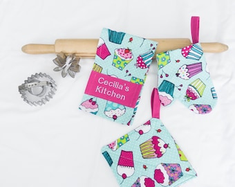 May include: A kitchen set featuring a rolling pin, a cookie cutter, and a set of cupcake-themed accessories. The accessories include a potholder, an oven mitt, and a dish towel, all with a light blue background and pink accents. The dish towel has the text "Cecilia's Kitchen".